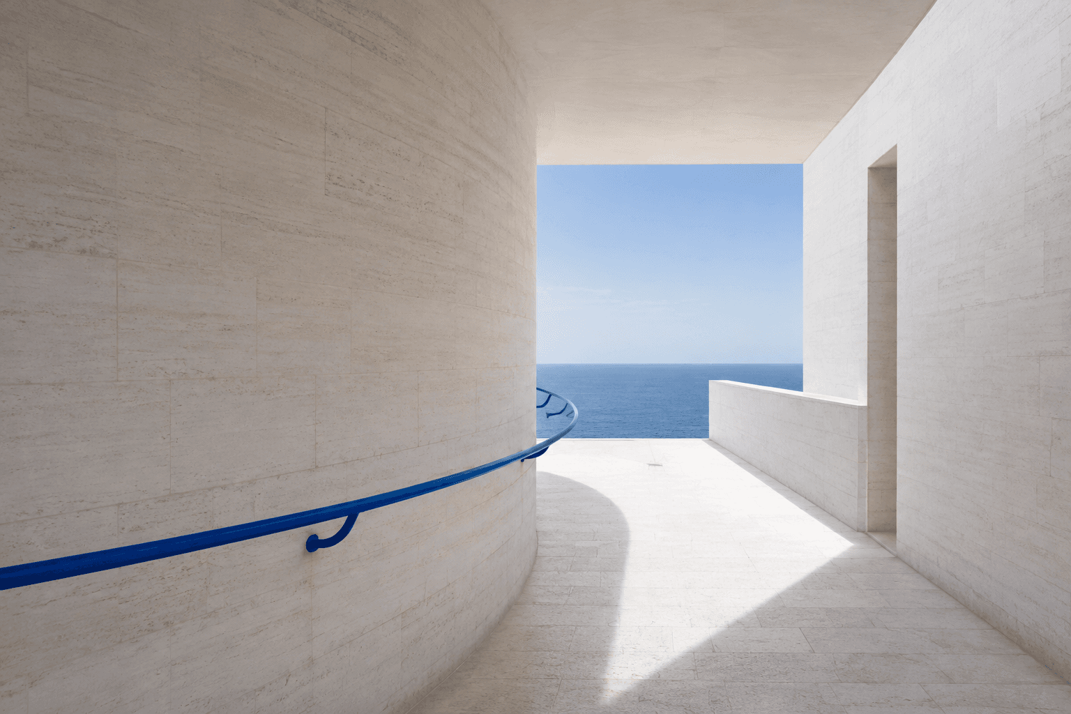Conceptual architecture: a pale stone corridor and blue handrail opening toward the sea